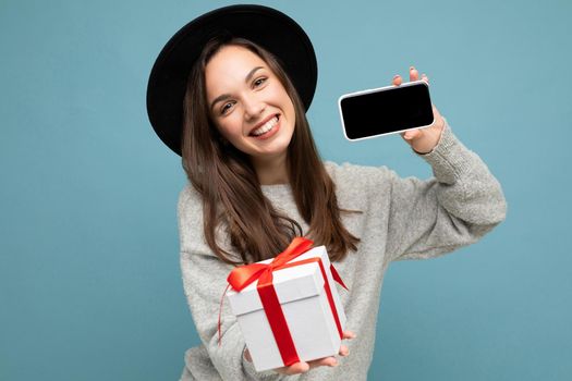 Shot of pretty smiling positive young brunet female person isolated over blue background wall wearing stylish black hat and grey sweater holding gift box showing smartphone screen display for mockup and looking at camera.