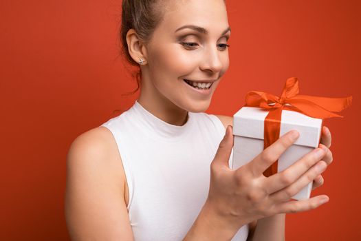 Closeup photo shot of beautiful happy smiling adult blonde woman isolated over red background wall wearing white top holding gift box and looking at white box with red ribbon.