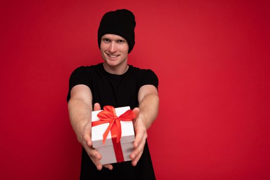 Shot of handsome happy smiling young man isolated over red background wall wearing black hat and black t-shirt holding white gift box with red ribbon and looking at camera.