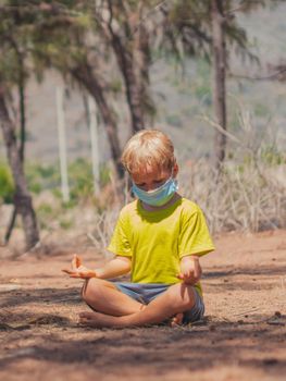 Calm little male boy sit meditation practice yoga forest park fresh air, wear COVID virus protective face mask. Young generation child rest relieve negative emotions close eye lotus pose mindfulness.