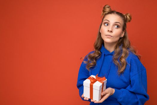 Shot of charming positive young blonde female person isolated over red background wall wearing blue trendy hoodie holding gift box and looking to the side and thinking. Empty space