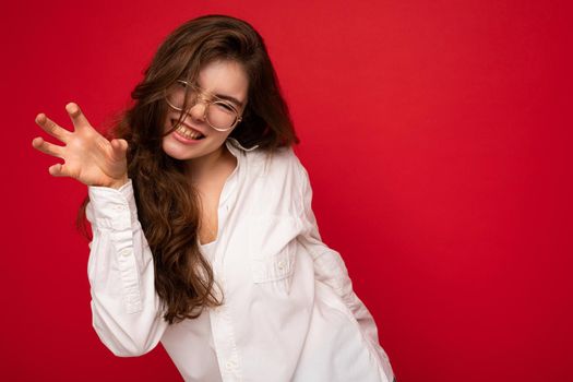 Portrait of young emotional positive sexy beautiful brunette woman with sincere emotions wearing casual white shirt and optical glasses isolated on red background with copy space and making cat claws and growling like animal.