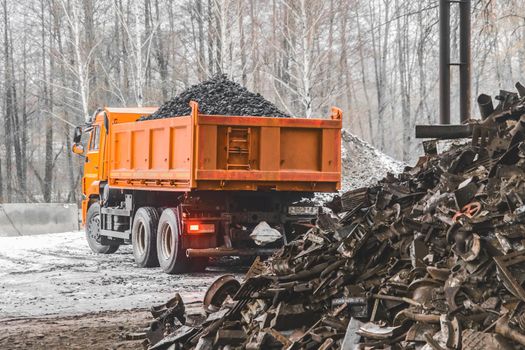 A dump truck in an industrial area or on a construction site in winter unloads coking coal from the body, next to a pile of metal waste and garbage.