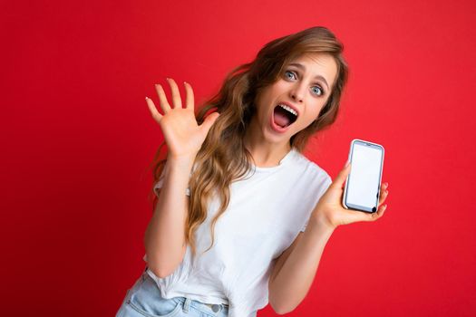 Amazed Shocked beautiful smiling young blonde woman good looking wearing white t-shirt standing isolated on red background with copy space holding phone showing smartphone in hand with empty screen display for mockup looking at camera showing open palm.