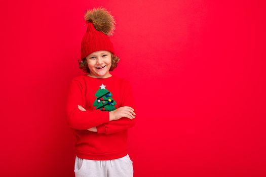 cool blond kid in warm hat and sweater with christmas tree on red background fooling around, christmas concept.