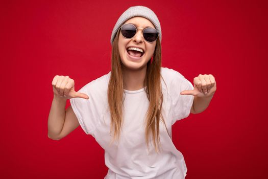 Photo of young emotional positive happy attractive dark blonde woman with sincere emotions wearing casual white t-shirt with empty space for mockup gray hat and sunglasses isolated on red background with copy space and having fun. Good mood concept.