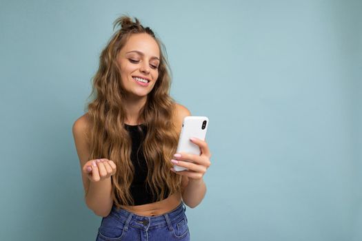 Positive smiling beautiful good looking young woman wearing casual stylish outfit poising isolated on background with empty space holding in hand and using mobile phone messaging sms looking at gadjet.