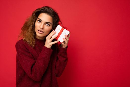 Beautiful thoughtful young brunette curly female person isolated over red background wall wearing red sweater holding gift box looking at camera. Free space