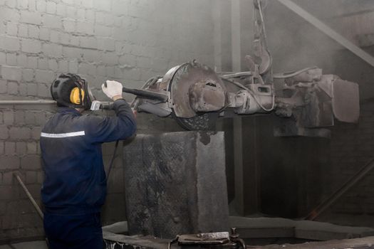 A man in work clothes, and a respirator drives heavy equipment with grinding stone to clean and prepare cast iron concrete tubing in a dirty shop of an industrial plant.