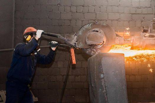 A male worker in a protective helmet, respirator, overalls manages heavy grinding equipment for cast iron concrete tubing with flying sparks in the workshop of an industrial plant.