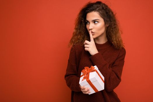 Photo shot of beautiful positive young brunette curly woman isolated over red background wall wearing red sweater holding gift box looking to the side and showing shh gesture. Copy space