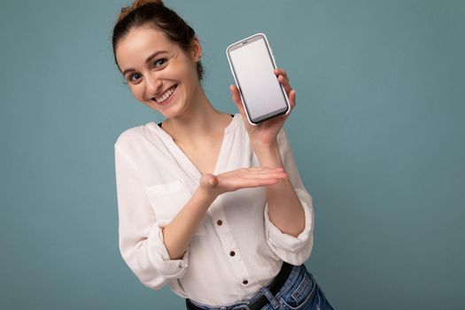 Beautiful positive smiling young blonde curly woman wearing casual white shirt and isolated over blue background wall holding phone and showing smartphone with empty dcreen for mockup looking at camera.