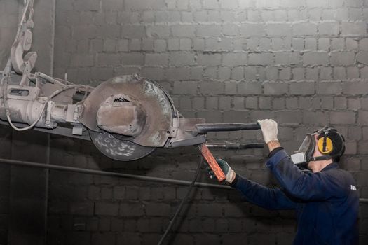 A man in work clothes and a respirator, controls heavy equipment with grinding stone to clean reinforced concrete structures in a dirty shop of an industrial plant.
