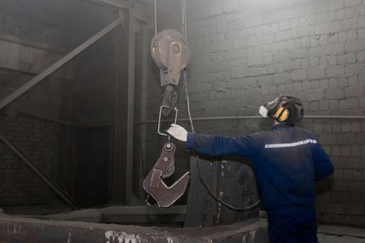 A man in a working jumpsuit and respirator takes a hook with his hand an industrial lifting mechanism in the factory workshop.