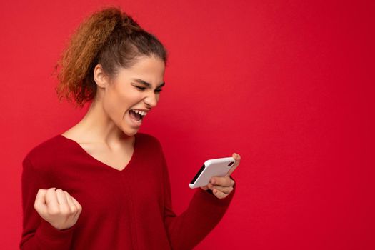 Young emotional woman wearing dark red sweater isolated over red background holding smartphone having fun and showing yes gesture looking at mobile phone screen.