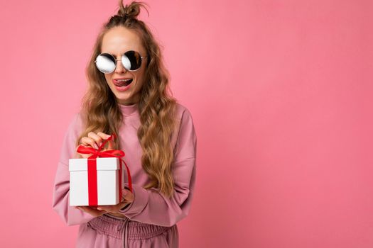 Shot of beautiful surprised positive young blonde curly woman isolated over pink background wall wearing pink sport clothes and sunglasses holding gift box and unboxing surprise. Copy space