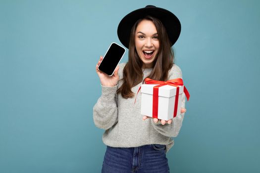 Fascinating happy joyful young brunette woman isolated over blue background wall wearing stylish black hat and grey sweater holding gift box showing mobile phone display for mockup and looking at camera.