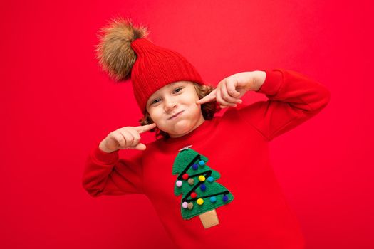 cool teenager in a red Christmas sweater fooling around against the background of a red wall, a warm hat and a sweater with a Christmas tree.