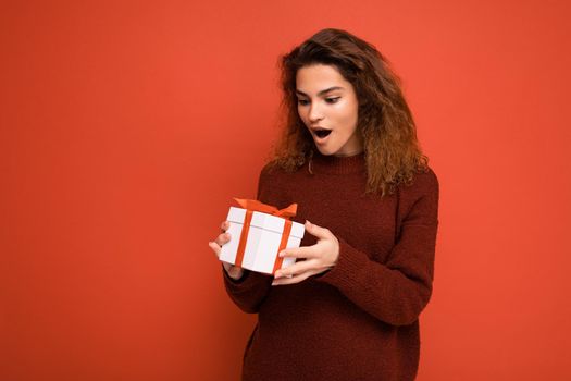 Attractive positive surprised young brunette curly woman isolated over red background wall wearing white sweater holding gift box and unboxing present looking at box with red ribbon. Copy space, mockup