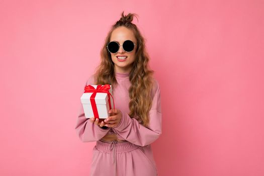 Shot of attractive positive smiling young blonde woman isolated over colourful background wall wearing everyday trendy outfit holding gift box and looking at camera.