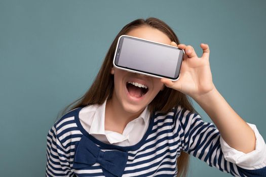 Photo of beautiful smiling happy teenager good looking wearing casual stylish outfit standing isolated on background with copy space holding smartphone showing phone in hand with empty screen display for mockup.