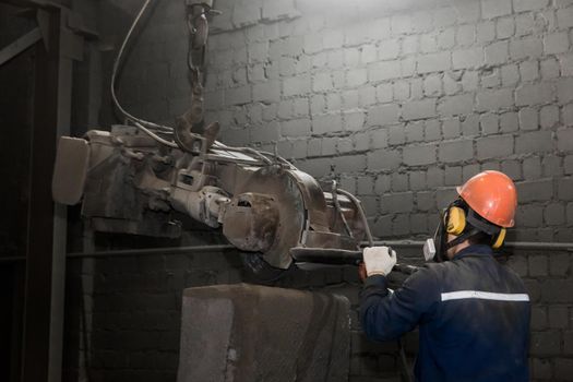 A working man in a protective helmet, respirator and overalls controls heavy grinding equipment for cleaning cast iron reinforced concrete structures and materials in the workshop of an industrial plant.