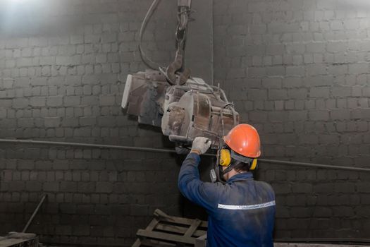 A working man in overalls, a protective helmet and a respirator controls heavy grinding equipment in the workshop of an industrial plant.