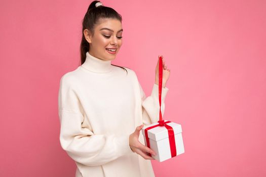 Attractive positive surprised young brunette woman isolated over pink background wall wearing white sweater holding gift box and unboxing present looking at box.