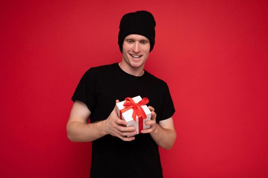 Shot of handsome happy young man isolated over red background wall wearing black hat and black t-shirt holding white gift box with red ribbon and looking at camera.