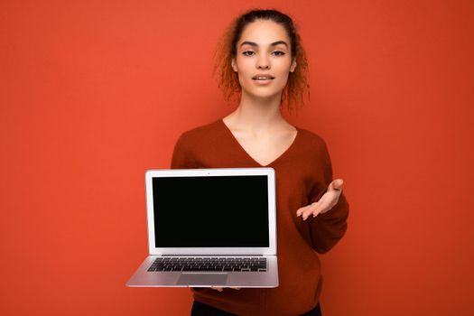 Beautiful charming fascinating young curly dark blond woman wearing red sweater standing isolated over red wall background holding computer laptop with empty copy space mock up pointing to the camera looking at camera and thinking about something with self-confidence.