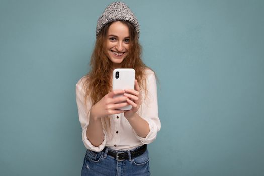 photo of attractive positive young blonde curly woman wearing casual white shirt and grey hat isolated over blue background wall holding phone and using communicating online looking at camera.