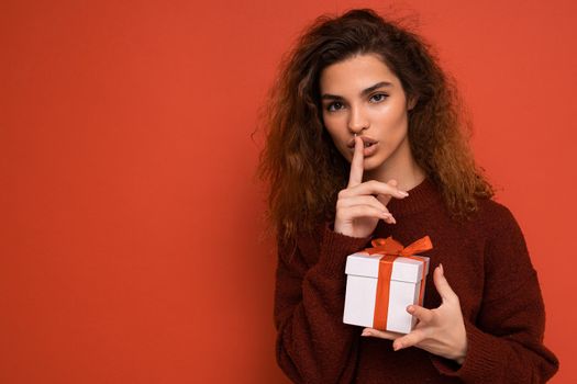 Shot of pretty young brunette curly woman isolated over red background wall wearing red sweater holding gift box looking at camera and showing shh gesture and meaning be quiet. Copy space, mockup