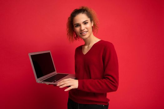 Close-up portrait of beautiful asking and surprised dark blond woman holding laptop computer looking at camera typing on keyboard wearing red sweater isolated over red wall background.