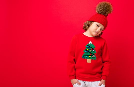 cool blond kid in warm hat and sweater with christmas tree on red background fooling around, christmas concept.