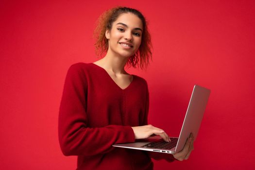Close-up portrait of beautiful smiling fascinating charming dark blond woman holding laptop computer looking at camera typing on keyboard wearing red sweater isolated over red wall background.