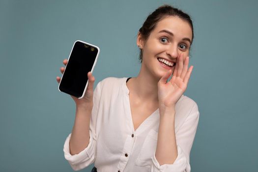 Beautiful positive young blonde curly woman wearing casual white shirt and isolated over blue background wall holding phone and showing smartphone with empty dcreen for mockup looking at camera and saying secret.