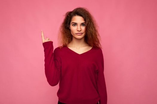 Photo of young serious beautiful brunette wavy woman with sincere emotions wearing casual pink jersey isolated over pink background with copy space and pointing at free space for mockup.