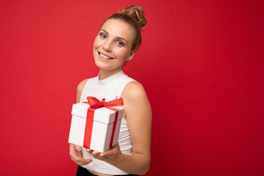 Photo shot of beautiful positive smiling young blonde woman isolated over red background wall wearing white top holding gift box and looking at camera. Copy space, mockup