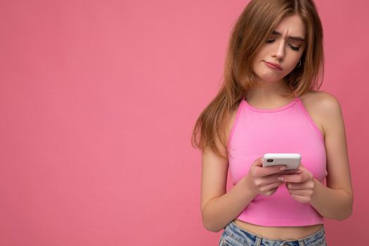 Closeup portrait of beautiful thoughtful young blonde woman wearing pink top poising isolated on pink background with empty space holding in hand and using mobile phone surfing on the internet online looking at gadjet display and having doubts. Copy space