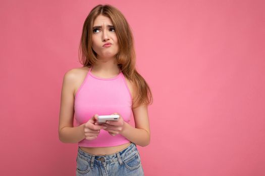 Beautiful thoughtful young blonde woman wearing pink top poising isolated on pink background with empty space holding in hand and using mobile phone surfing on the internet online looking to the side and having doubts. copy space