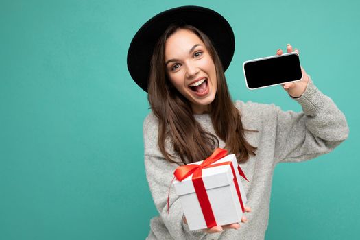 Photo of beautiful happy joyful young brunette woman isolated over blue background wall wearing stylish black hat and grey sweater holding gift box showing mobile phone screen display for mockup and looking at camera.