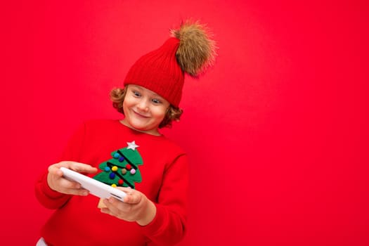 a young man with a smartphone in his hands against the background of a red wall, in a knitted hat and a sweater with a Christmas tree