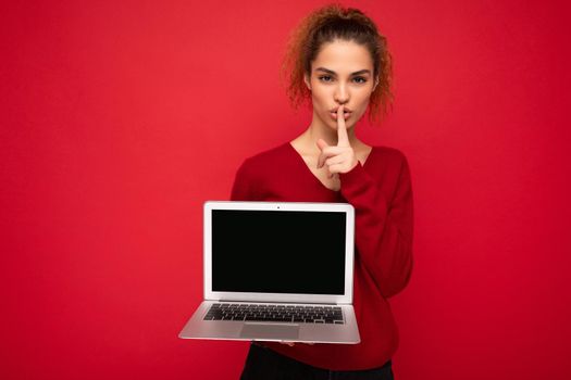 Close-up portrait of beautiful dark blond curly young woman holding laptop computer looking at camera holding finger close to mouth and lips showing gesture shhh and saying hush be quiet wearing red sweater isolated over red wall background. Mock up copy empty space.