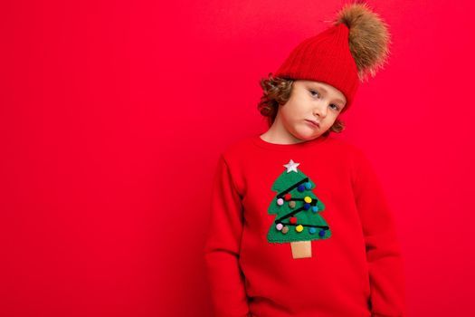 cool teenager in a red Christmas sweater fooling around against the background of a red wall, a warm hat and a sweater with a Christmas tree.
