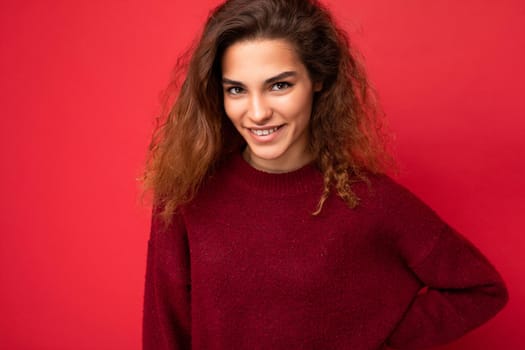 Young beautiful curly brunette woman with sincere emotions poising isolated on background wall with copy space wearing casual dark red sweater. Fun and joy concept.