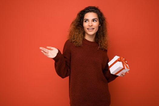 Beautiful happy young brunette woman isolated over colourful background wall wearing stylish casual clothes holding gift box and looking to the side.