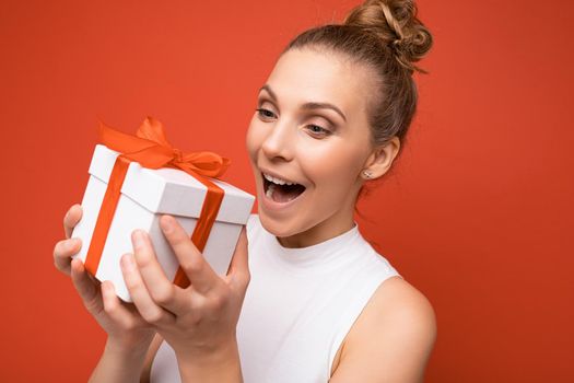 Closeup photo shot of attractive positive surprised amazed young blond woman isolated over colourful background wall wearing trendy outfit look holding gift box and looking at present box with red ribbon.