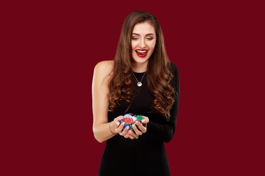 Casino, gambling, poker, people and entertainment concept - woman poker player in black dress with chips in hands on red background. Studio shot
