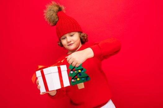 a cool boy with curls on a red wall background in a sweater with a Christmas tree holding a gift box in his hands.
