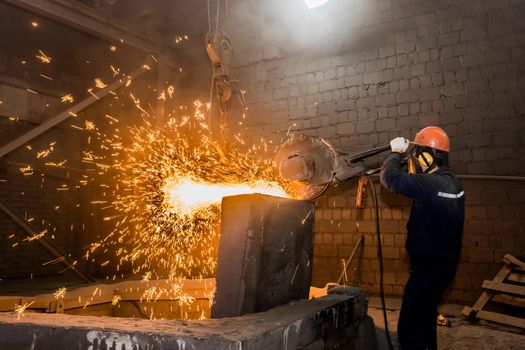 A male worker in a protective helmet, respirator, overalls manages heavy grinding equipment for cast iron concrete tubing with flying sparks in the workshop of an industrial plant.